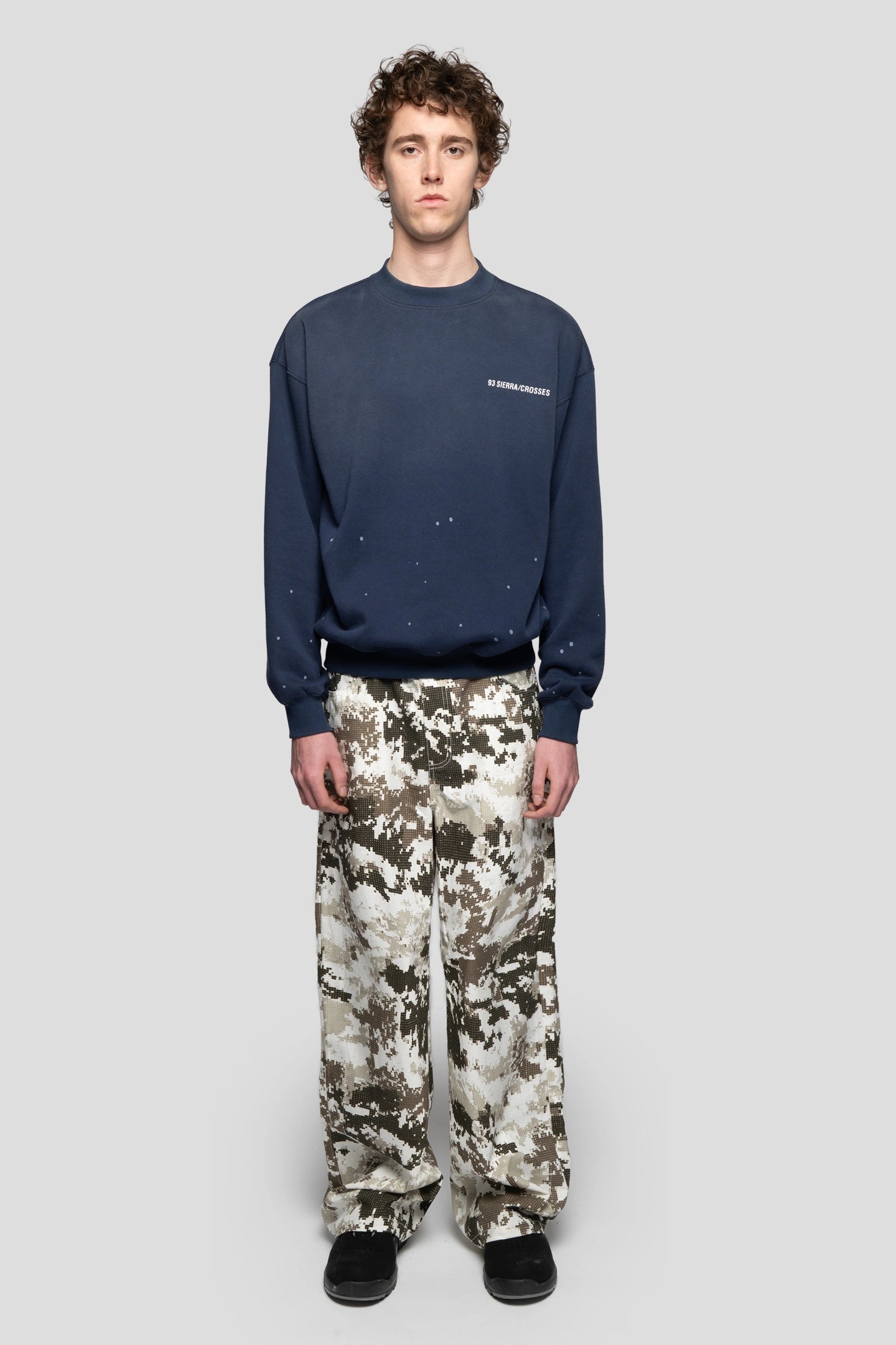 Model wearing a navy blue crewneck with a paint splattered effect  and the logo of the brand with black, brown and white camo pants